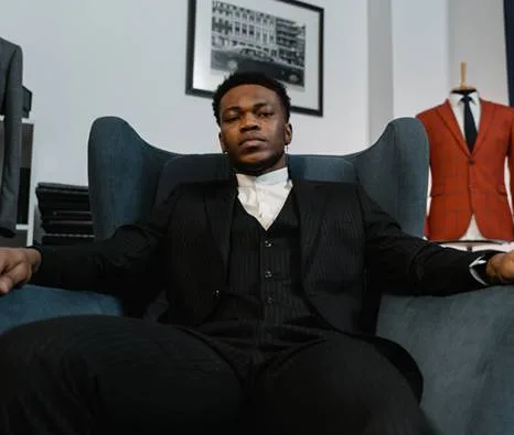 Man in black three-piece suit sitting confidently on armchair indoors